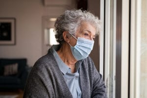 Lonely old woman wearing surgical mask. Sad senior lady wearing face protective medical mask and looking through the window. Alone depressed woman stay at home during quarantine due to the coronavirus