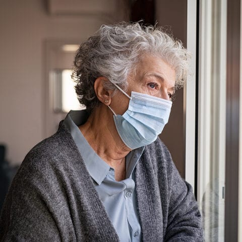 Lonely old woman wearing surgical mask. Sad senior lady wearing face protective medical mask and looking through the window. Alone depressed woman stay at home during quarantine due to the coronavirus