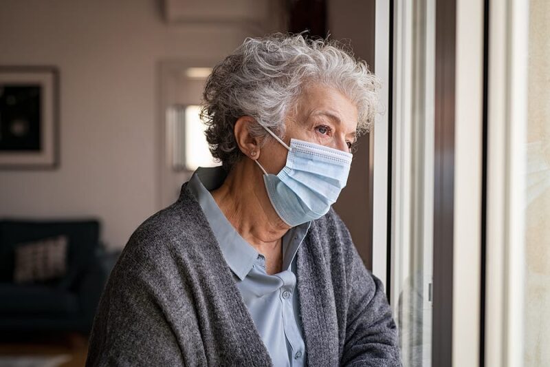 Lonely old woman wearing surgical mask. Sad senior lady wearing face protective medical mask and looking through the window. Alone depressed woman stay at home during quarantine due to the coronavirus