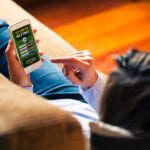 Woman holding a mobile phone to visit a sports betting website while lies down at home.