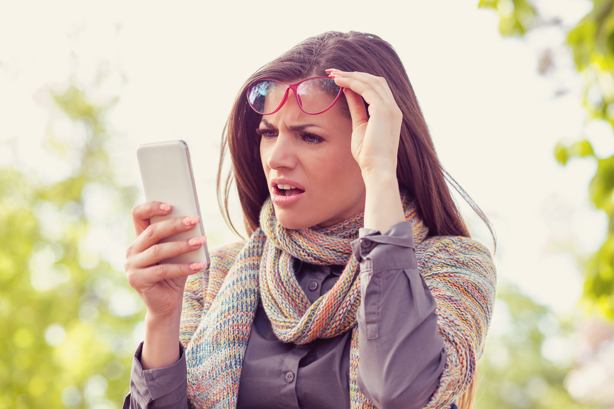 Annoyed upset woman in glasses looking at her smart phone with frustration while walking on a street. Photo by Chiosea