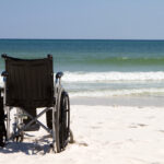 Empty wheelchair sits vacant on a beach of sand with ocean waves and surf in the background.