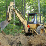 Operator using back hoe for excavating in forest.
