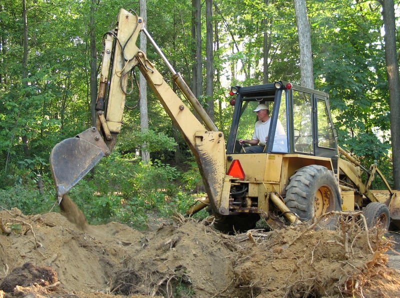 Operator using back hoe for excavating in forest.