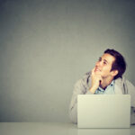 Young man in office, sitting at desk with laptop daydreaming, smiling