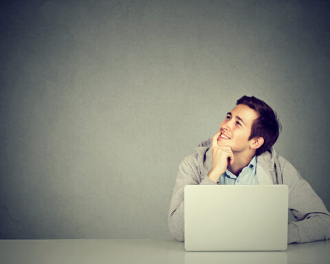 Young man in office, sitting at desk with laptop daydreaming, smiling
