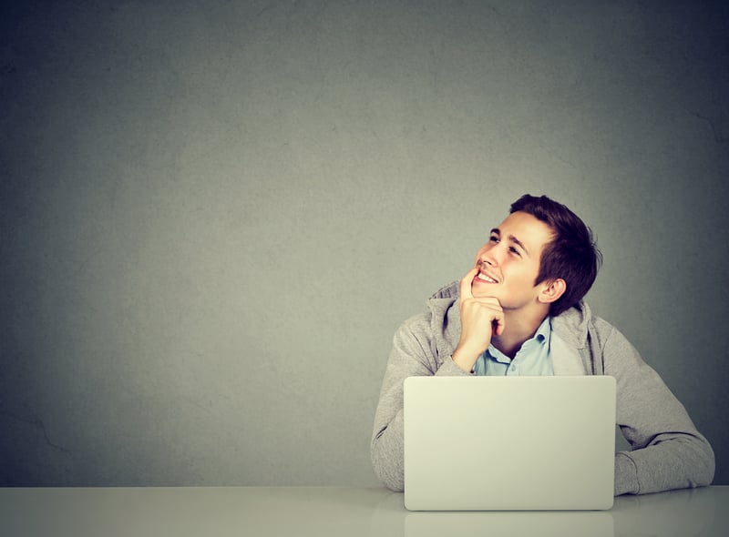Young man in office, sitting at desk with laptop daydreaming, smiling