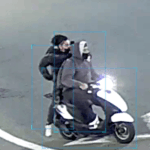 Two people on a white and blue scooter: a front rider in a gray hoodie, a second rider in a dark jacket seated behind.