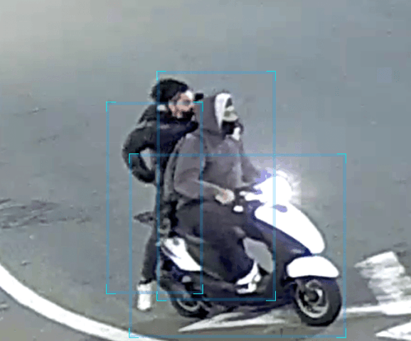 Two people on a white and blue scooter: a front rider in a gray hoodie, a second rider in a dark jacket seated behind.