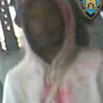Surveillance image of a hooded person with face blurred, wearing a light-colored jacket and pink shirt, with a police badge overlay in the corner.
