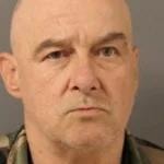 Mugshot-style portrait of a bald, middle-aged man with a stern expression wearing camouflage clothing.