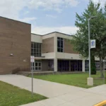 Exterior view of a brick school or college building with large glass windows, surrounded by a sidewalk, grass, and trees under a blue sky.