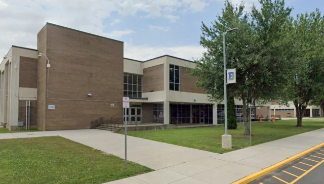Exterior view of a brick school or college building with large glass windows, surrounded by a sidewalk, grass, and trees under a blue sky.