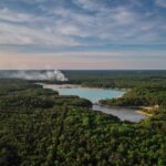 Aerial view of a dense forest with a smoke plume rising near a small lake.