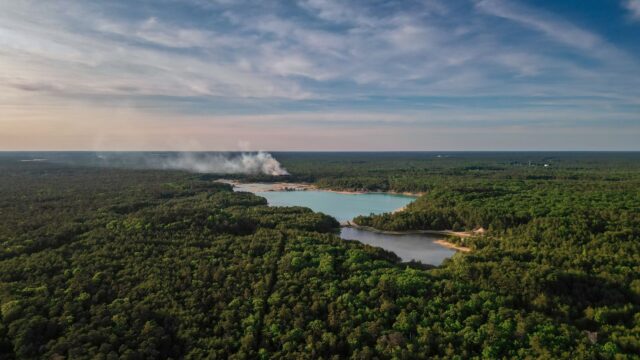 Aerial view of a dense forest with a smoke plume rising near a small lake.