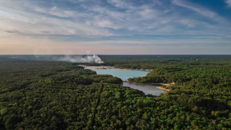 Aerial view of a dense forest with a smoke plume rising near a small lake.