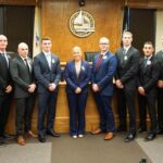 Group of ten people in formal attire posing in a wood-paneled council chamber with a city seal behind them on the wall.