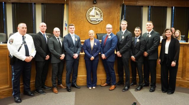 Group of ten people in formal attire posing in a wood-paneled council chamber with a city seal behind them on the wall.