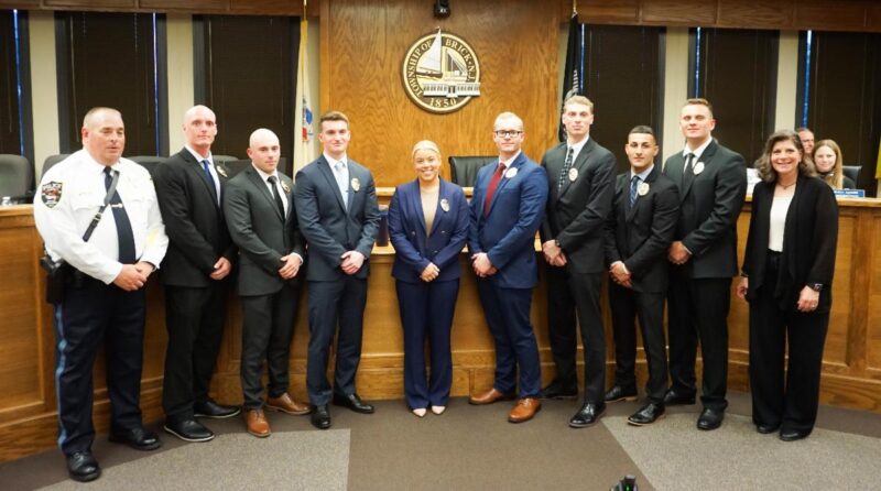 Group of ten people in formal attire posing in a wood-paneled council chamber with a city seal behind them on the wall.
