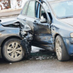 Two cars collide head-on, with crumpled hoods and shattered windshield debris in front of an urban street.