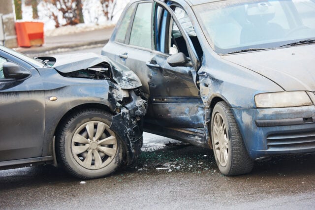 Two cars collide head-on, with crumpled hoods and shattered windshield debris in front of an urban street.