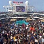 Crowd of passengers dancing around a pool on a cruise ship with multiple deck levels and a large central screen above.