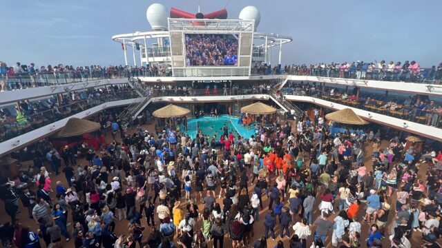 Crowd of passengers dancing around a pool on a cruise ship with multiple deck levels and a large central screen above.