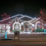 Two-story brick house glowing with Christmas lights along the roofline, wreath on the front, and lit trees in the yard at night in a festive display.