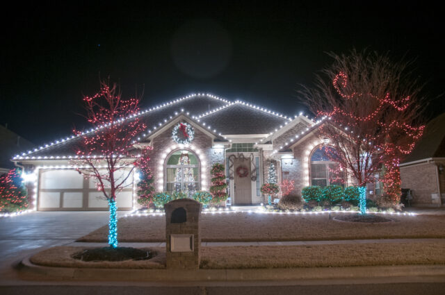 Two-story brick house glowing with christmas lights along the roofline, wreath on the front, and lit trees in the yard at night in a festive display.