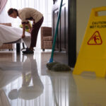 Housekeeper tucking a white sheet on a bed in a hotel room, with a yellow wet-floor sign in the foreground and a glossy floor reflecting the scene.