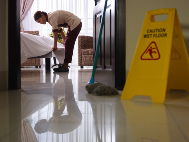 Housekeeper tucking a white sheet on a bed in a hotel room, with a yellow wet-floor sign in the foreground and a glossy floor reflecting the scene.