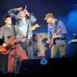 Three male musicians perform on a lit concert stage: a singer at the center singing into a microphone, a bassist on the left, and a guitarist on the right playing a sunburst bass guitar and an electric guitar respectively.