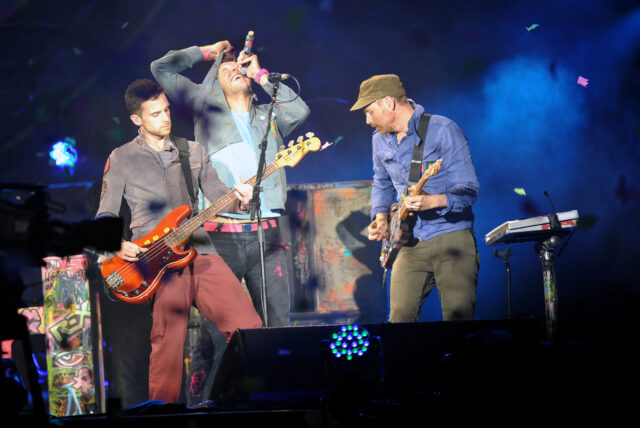 Three male musicians perform on a lit concert stage: a singer at the center singing into a microphone, a bassist on the left, and a guitarist on the right playing a sunburst bass guitar and an electric guitar respectively.