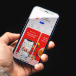 Hand holding a blue iPhone showing the DoorDash app with red promo panels and a food delivery interface on a dark background (informative app screenshot)