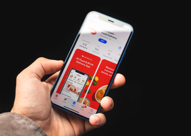 Hand holding a blue iphone showing the doordash app with red promo panels and a food delivery interface on a dark background (informative app screenshot)