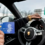Man wearing sunglasses driving a Porsche, holding a blue EBT card toward the camera.