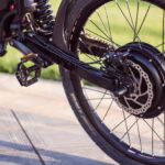 Close-up of a black bicycle's rear wheel, disc brake rotor, chain, and suspension against a grassy background in sunlight.