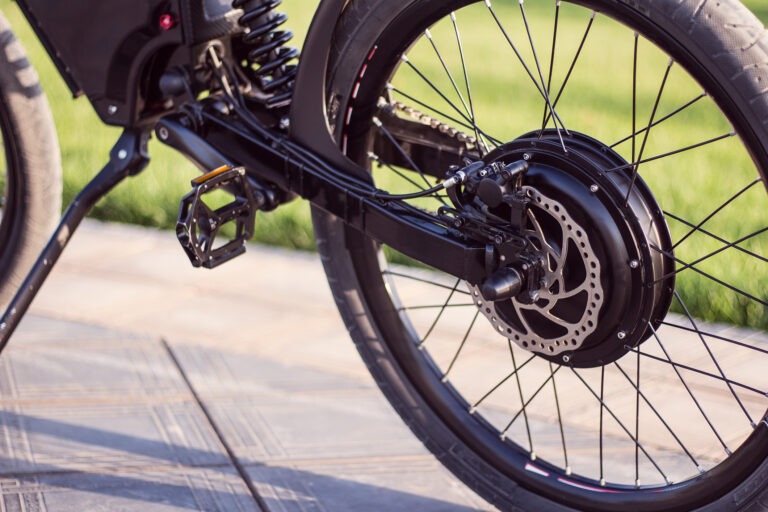 Close-up of a black bicycle's rear wheel, disc brake rotor, chain, and suspension against a grassy background in sunlight.