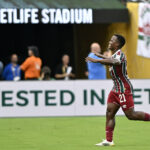 Soccer player in maroon and green stripes with number 21 runs with arms outstretched on a green field, METLIFE STADIUM sign above and spectators in the background.