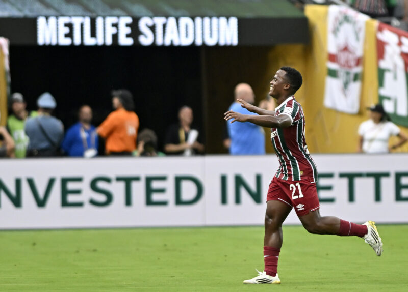 Soccer player in maroon and green stripes with number 21 runs with arms outstretched on a green field, metlife stadium sign above and spectators in the background.