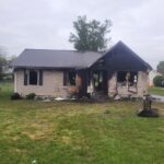 Front view of a house heavily damaged by fire, with charred siding and exposed beams in the yard outside the remains of the structure.