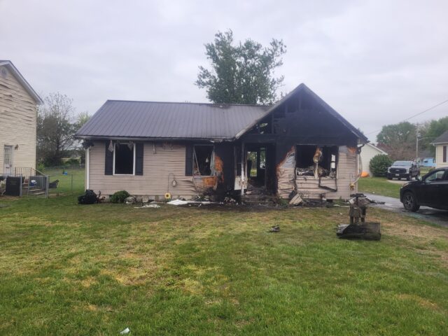 Front view of a house heavily damaged by fire, with charred siding and exposed beams in the yard outside the remains of the structure.