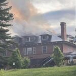 Brick multi-story building on fire with flames and thick smoke billowing from the roof; surrounding lawn and trees in the foreground.