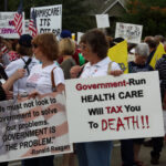 Crowd of protesters at a political rally holding signs and American flags; the prominent sign says, 'Government-Run HEALTH CARE Will TAX You To DEATH!!' A second sign quotes Ronald Reagan about government solving problems.