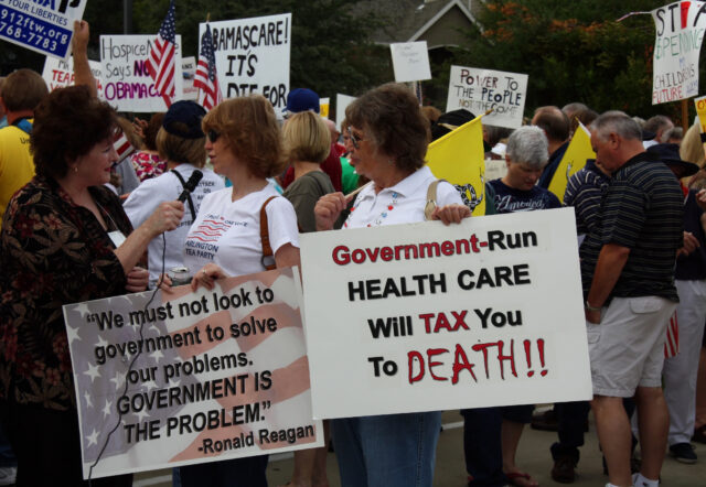 Crowd of protesters at a political rally holding signs and american flags; the prominent sign says, 'government-run health care will tax you to death!! ' a second sign quotes ronald reagan about government solving problems.