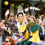 Two parade participants ride a float: one in a rainbow sequin dress and crown waves to the crowd while the other wears a leafy green costume with a feathered headpiece and beard of greenery, smiling at spectators.