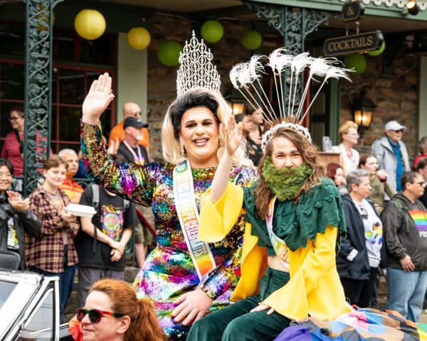 Two parade participants ride a float: one in a rainbow sequin dress and crown waves to the crowd while the other wears a leafy green costume with a feathered headpiece and beard of greenery, smiling at spectators.