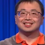Smiling man with glasses wearing an orange polo shirt against a blue curtain backdrop.