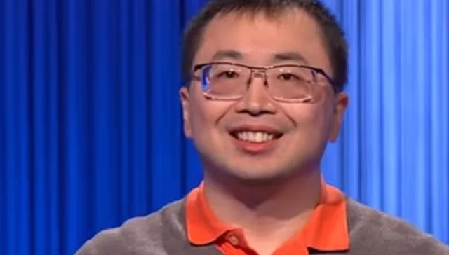 Smiling man with glasses wearing an orange polo shirt against a blue curtain backdrop.