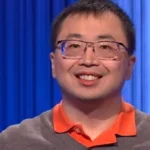 Smiling man with glasses in a gray sweater and orange polo, standing against blue backdrop curtains on a stage.